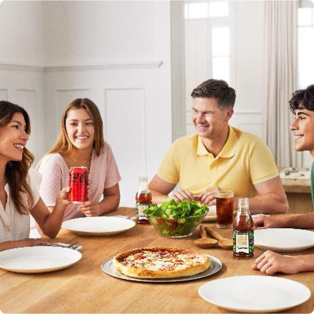 People sitting around a table with pizza at the center and someone holding a can of Coca-Cola